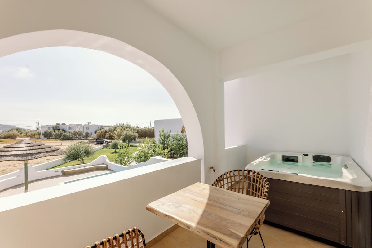 A balcony with a wooden table and chairs, a hot tub, and a view of a garden and buildings.
