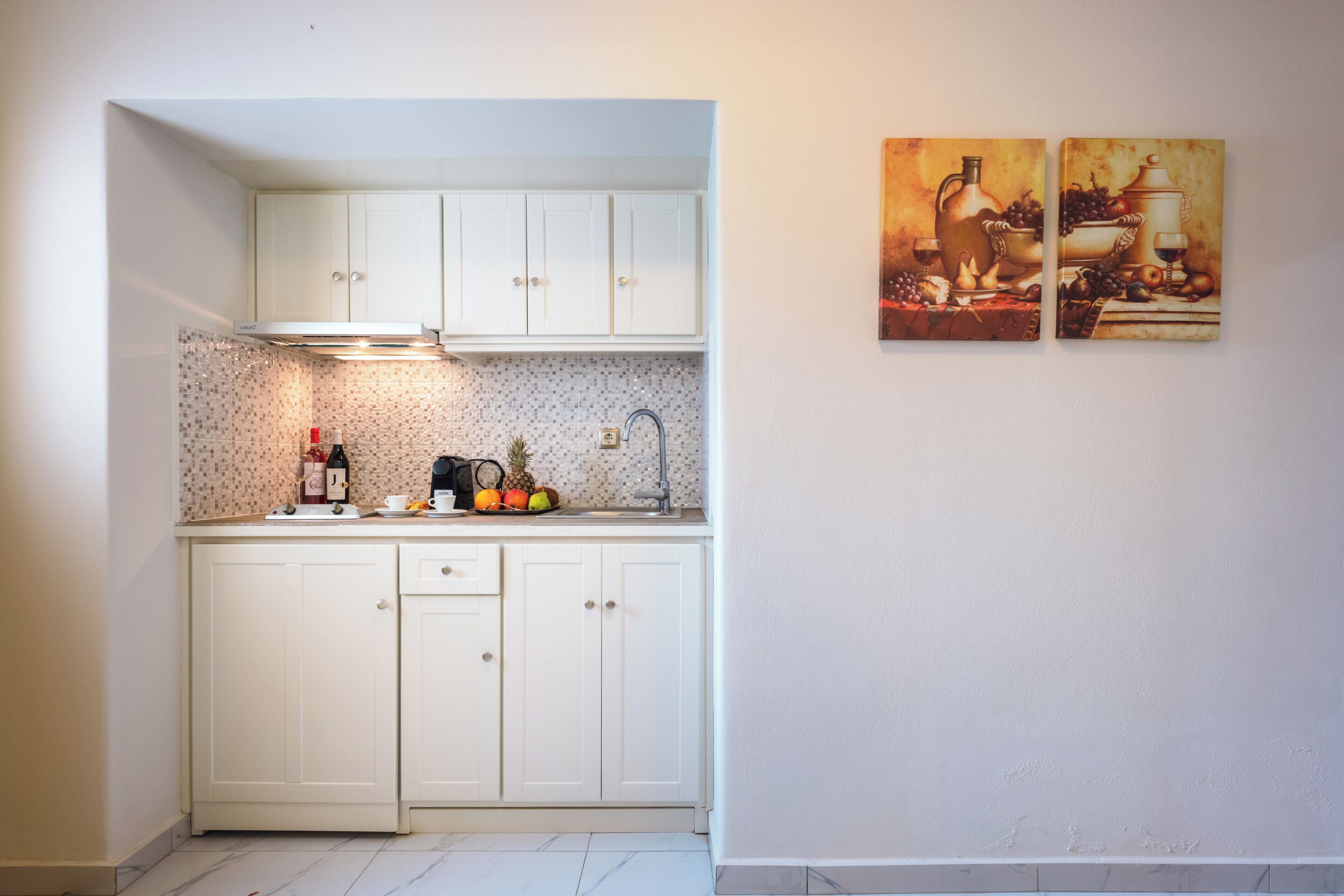 A compact kitchen with white cabinets, a countertop, and a backsplash. There are two wall-mounted paintings depicting still life scenes.