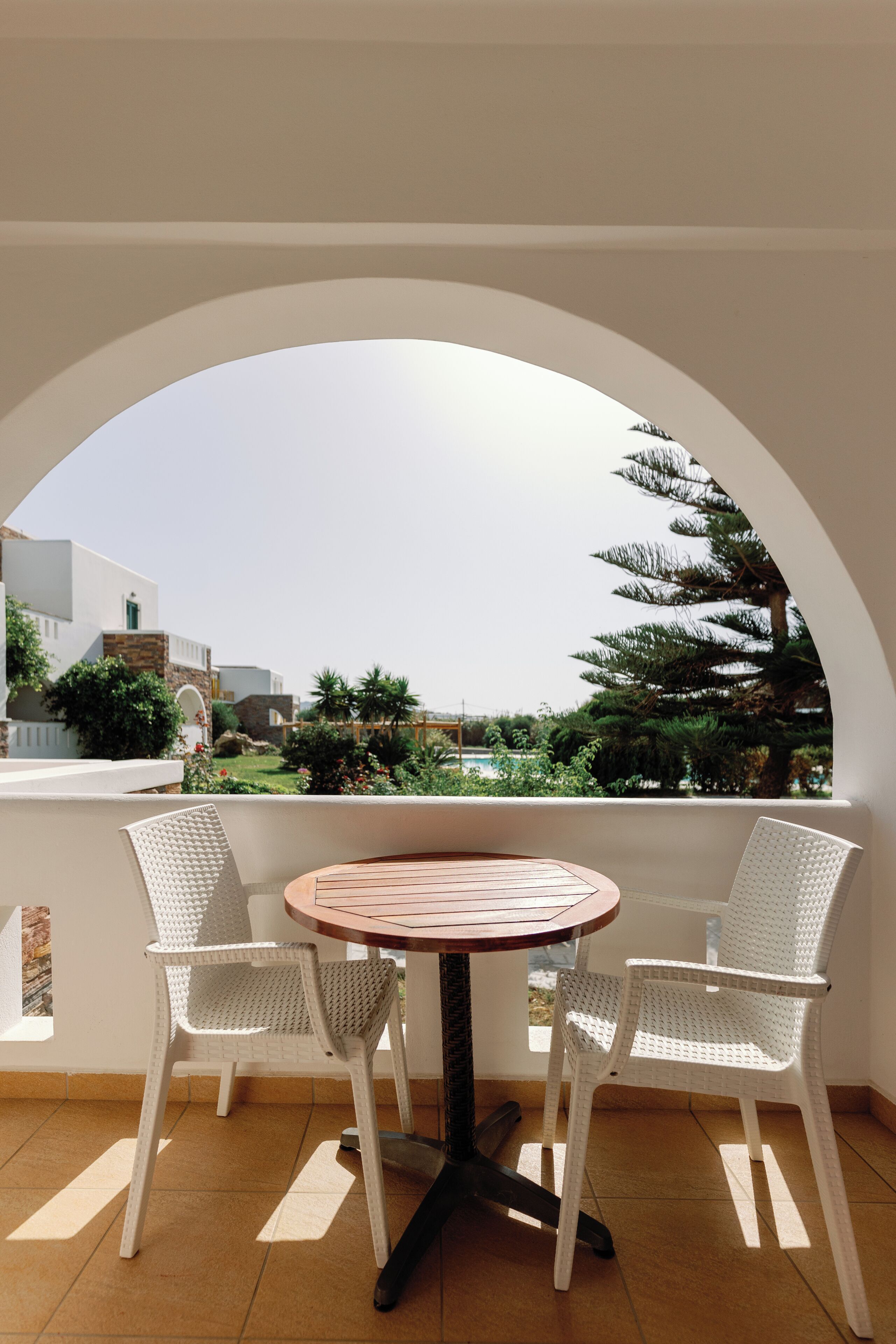 Un balcón con una mesa redonda y dos sillas blancas de mimbre, con vistas a un jardín y edificios.