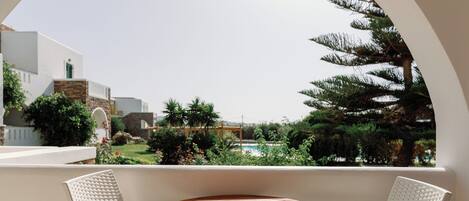 Un balcón con una mesa redonda y dos sillas blancas de mimbre, con vistas a un jardín y edificios.