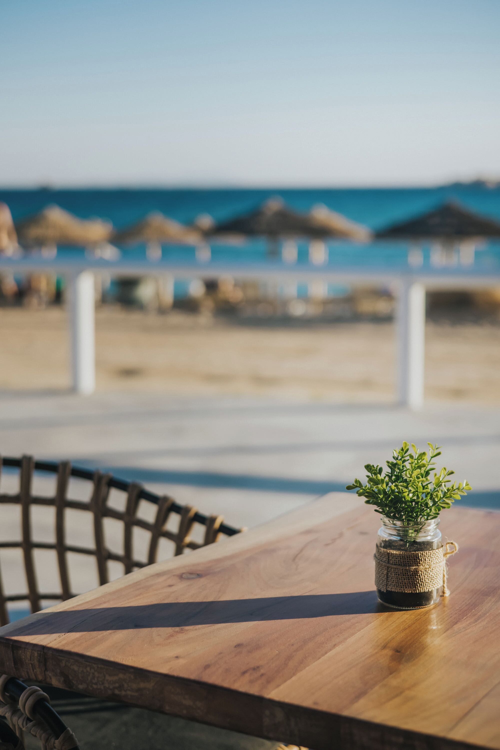 A wooden table with a small potted plant, a chair, and a view of the beach with umbrellas in the background.