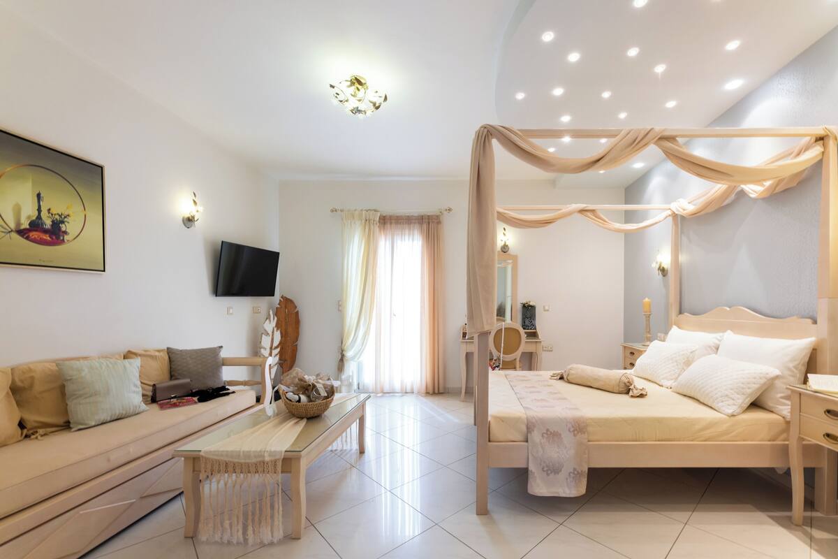A bedroom with a four-poster bed, a sofa, a TV, and a bedside table with a lamp.