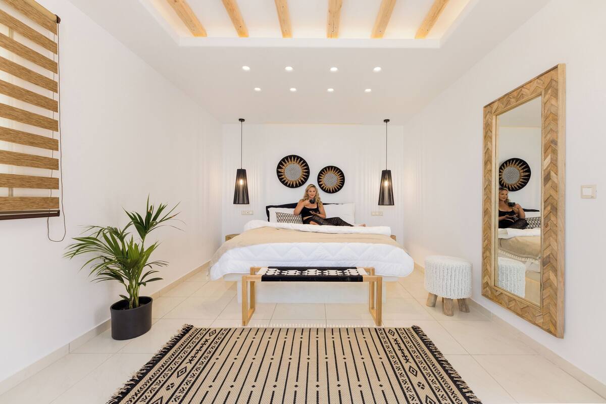 A modern bedroom with a large bed, bedside lamps, a potted plant, a bench, and a mirror.