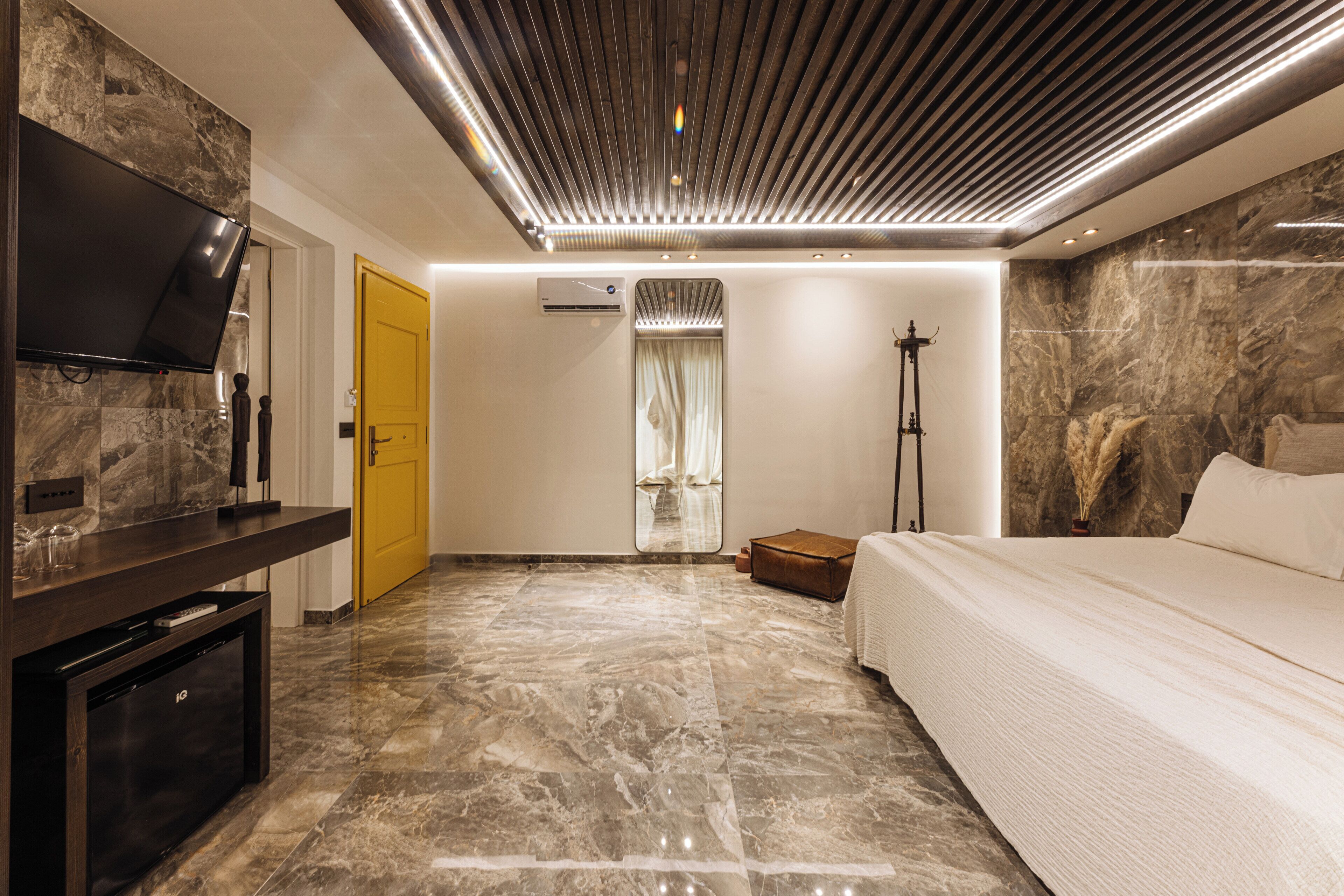A modern hotel room with a flat-screen TV, a bed, a seating area, and a bathroom with a mirror and towel rack.