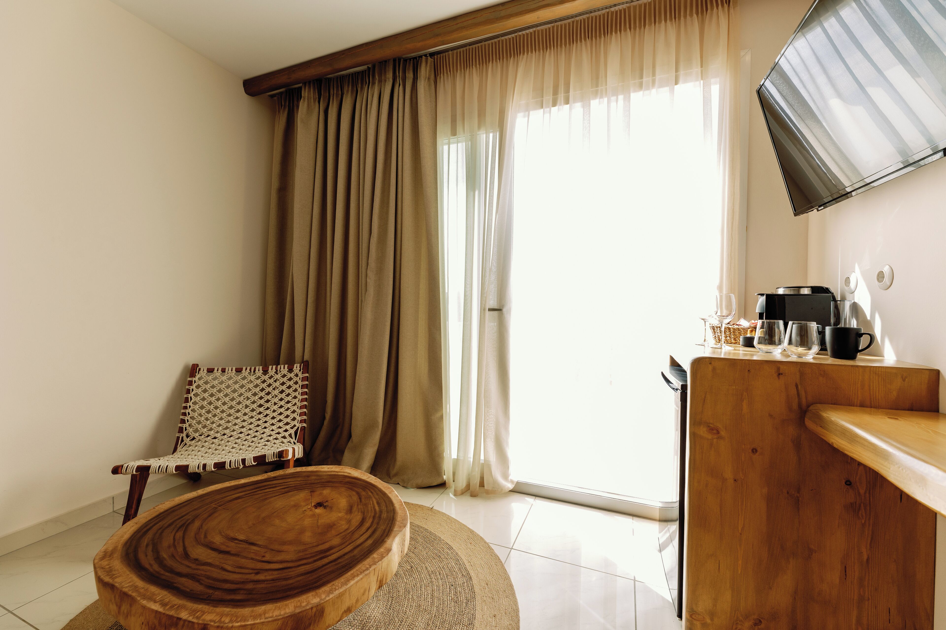 A room with a wooden table, a woven chair, a large window with curtains, and a coffee machine.