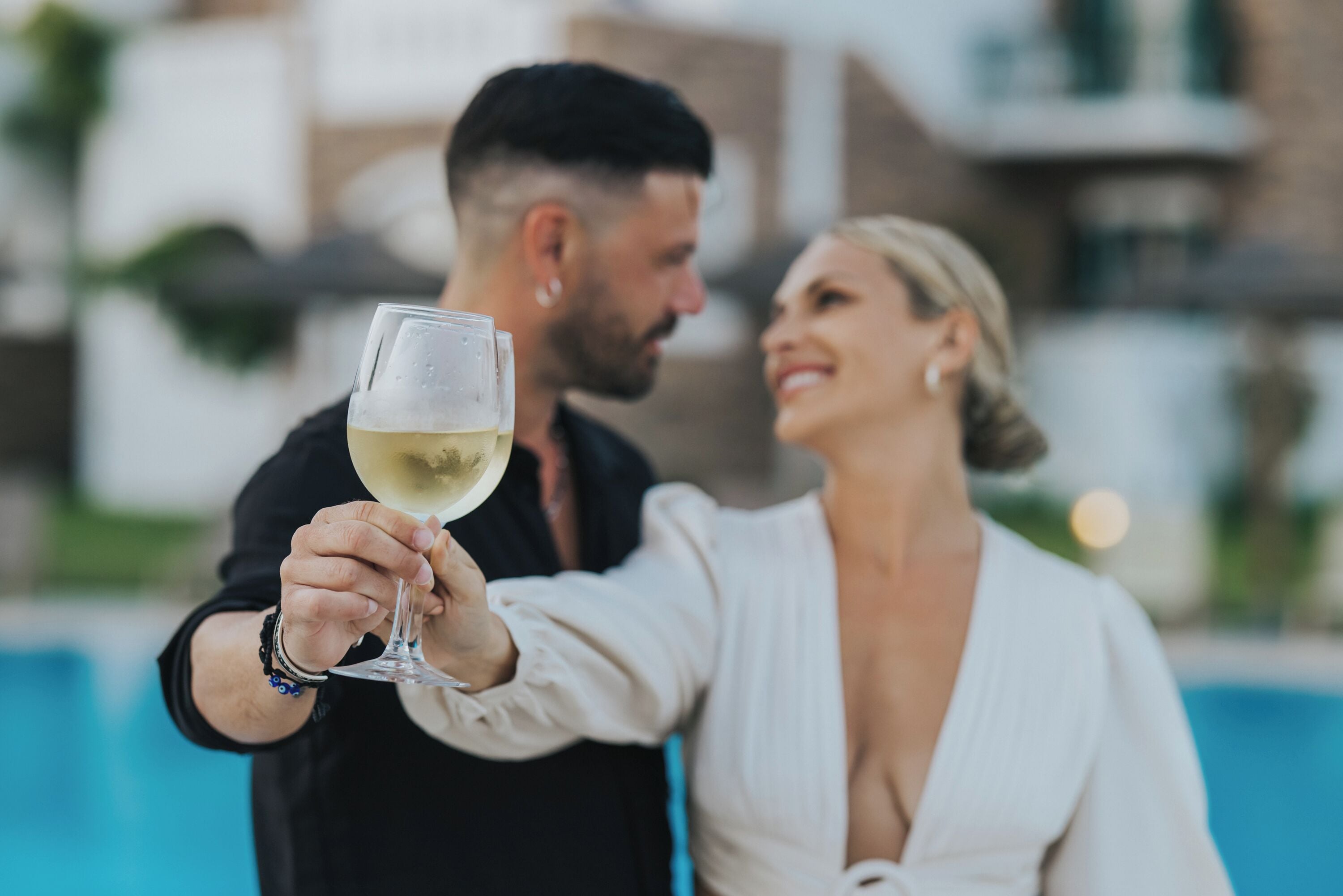 Un couple trinque avec un verre de vin blanc au bord d’une piscine.