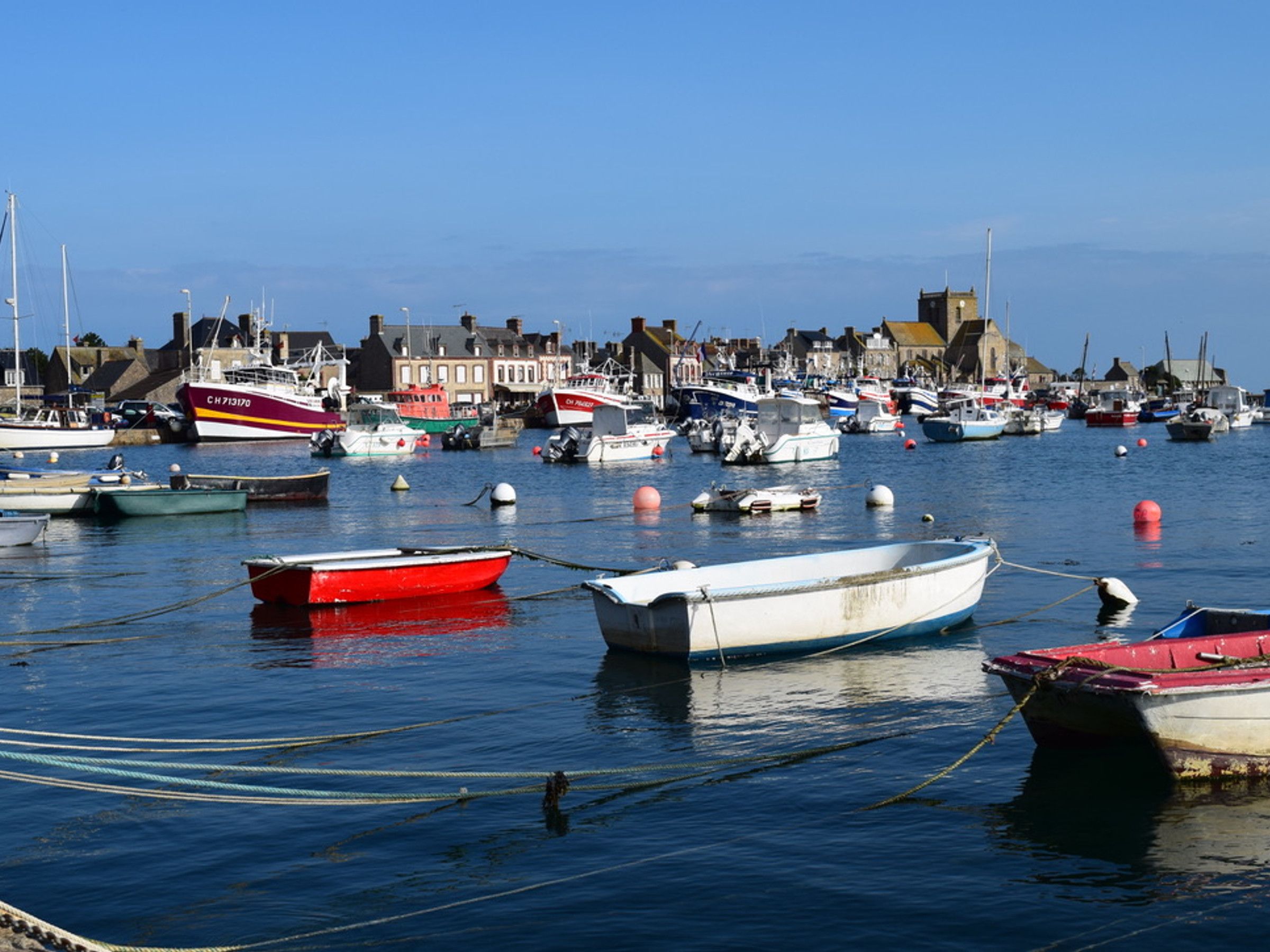A harbor with various boats docked, buildings in the background, and calm water.
