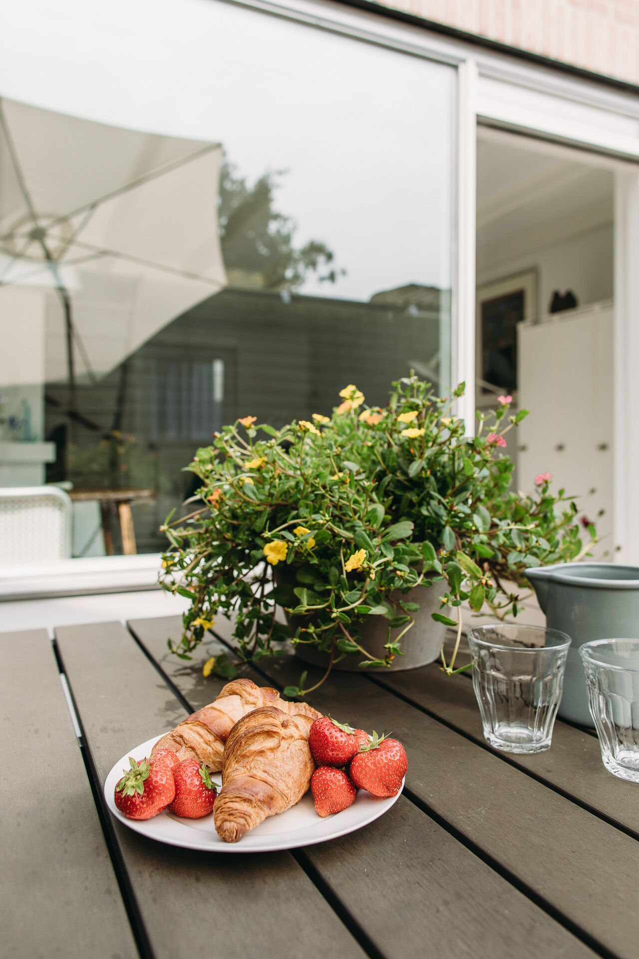 Une table avec des croissants et des fraises, une plante en pot et deux verres d’eau, le tout sur une terrasse.