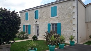 A two-story stone building with blue shutters, a small garden with potted plants, and a paved driveway.