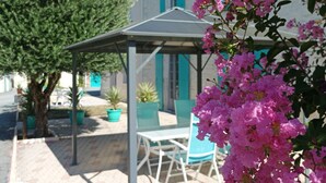 A covered outdoor seating area with a table and chairs, surrounded by potted plants and a tree.