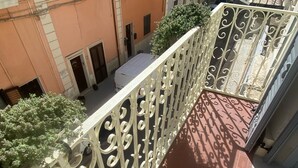 Ein Balkon mit aufwendiger Metallgeländerung, rotem Fliesenboden und Blick auf eine enge Straße mit parkenden Autos und Gebäuden.