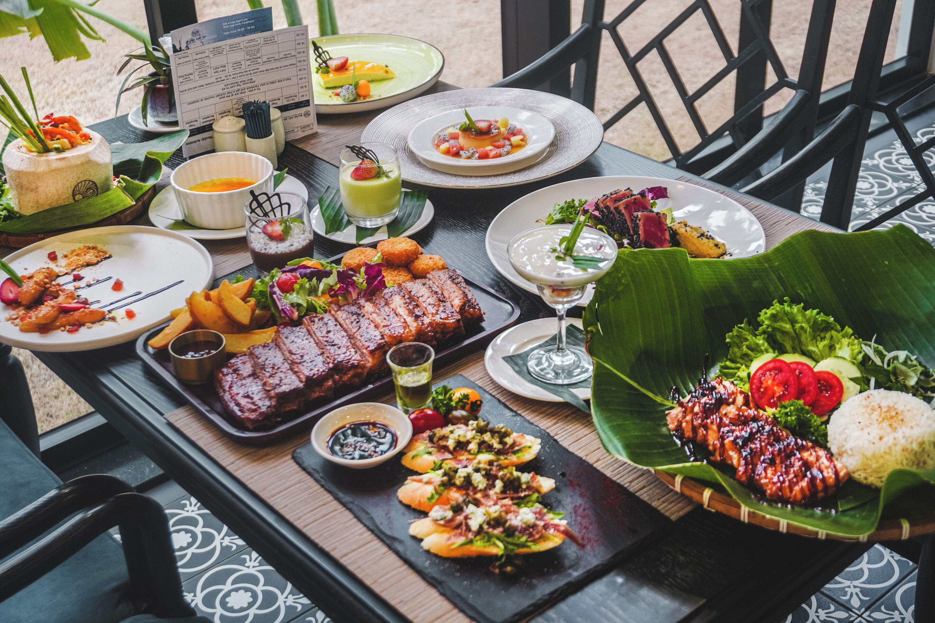 Una mesa dispuesta con diversos platos, incluyendo carnes a la parrilla, ensaladas y postres, sobre una mesa de madera con un menú y una planta al fondo.