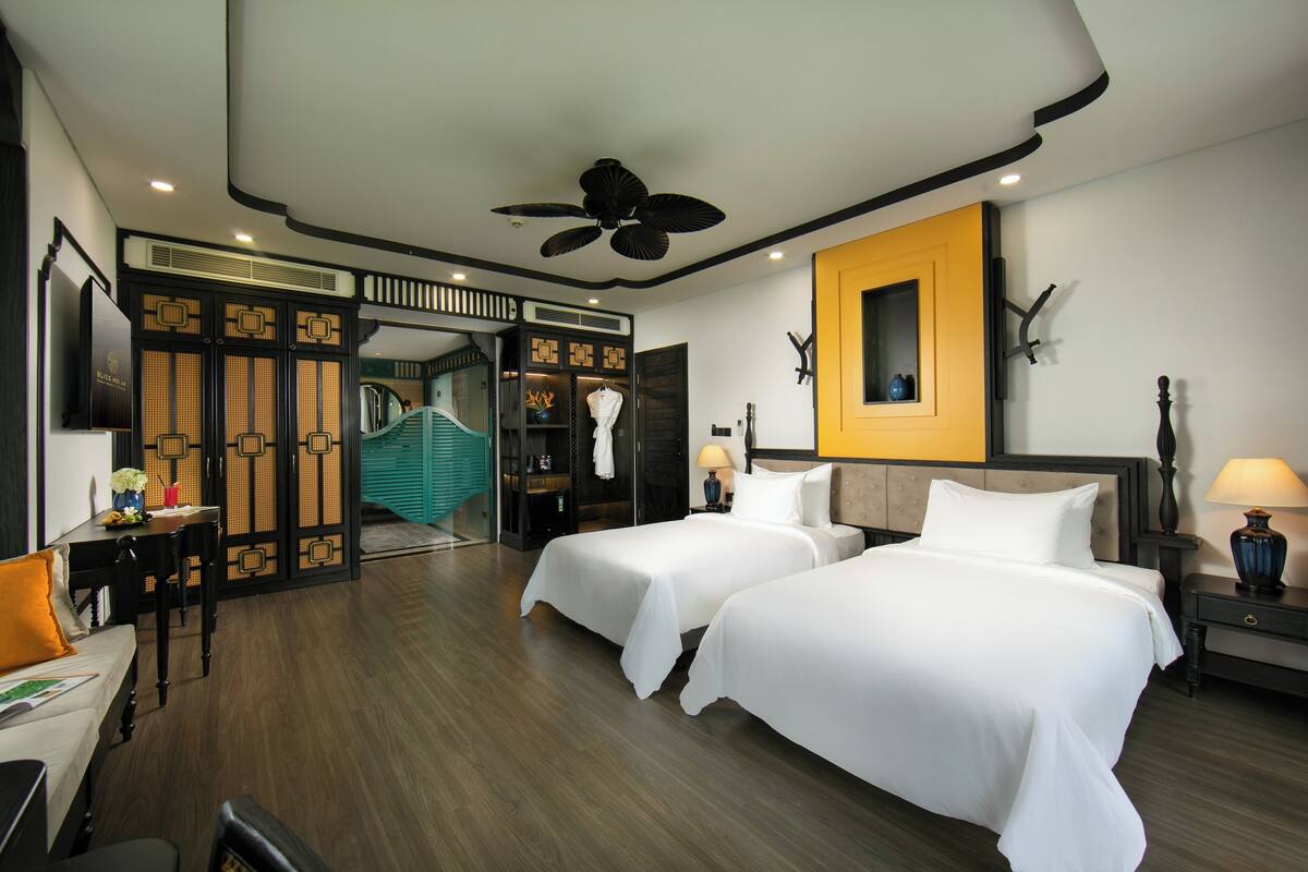 Une chambre d’hôtel avec deux lits, un ventilateur de plafond, une télévision, un bureau avec une chaise et une salle de bain avec douche et toilettes.