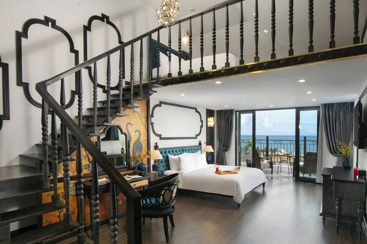 Une chambre spacieuse avec un grand lit, un bureau et une chaise, un escalier orné de balustrades travaillées et un balcon offrant une vue sur l’océan.