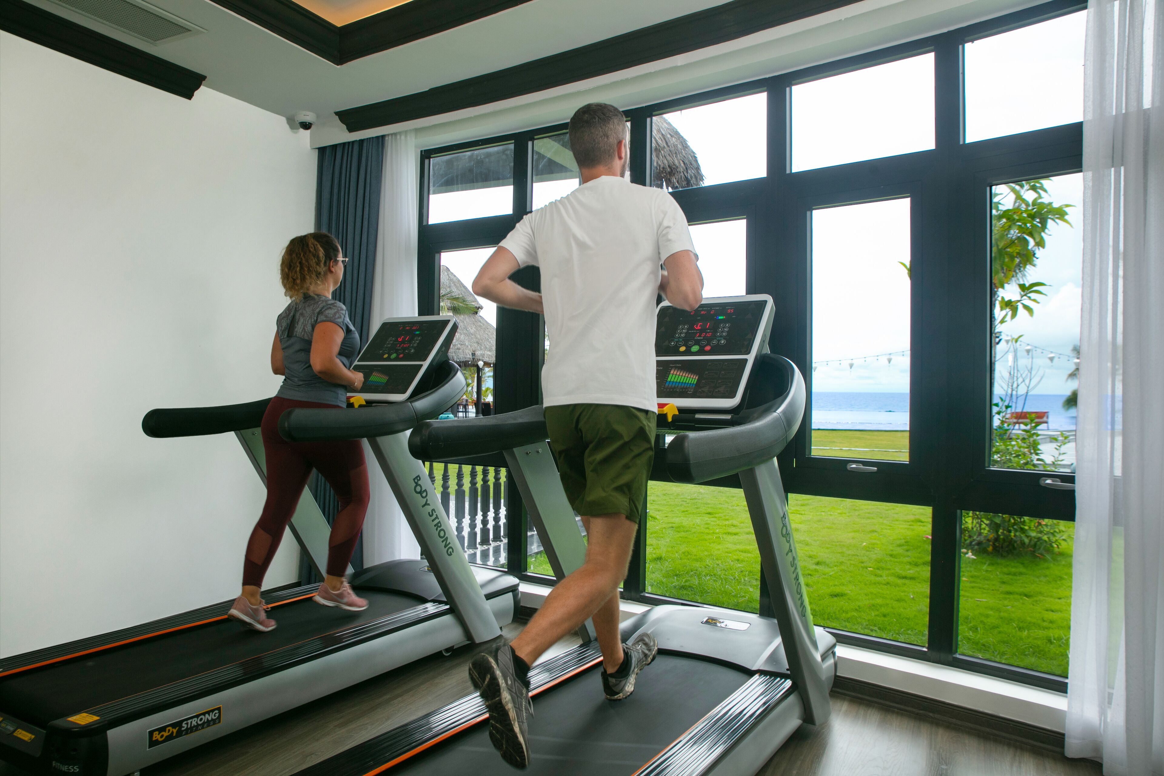 Two people running on treadmills in a room with large windows overlooking a grassy area.