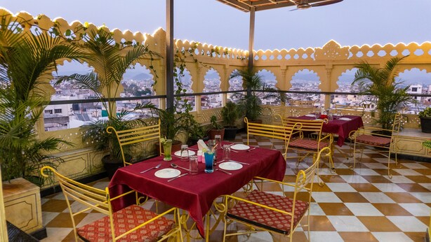 An outdoor dining area with checkered flooring, yellow metal chairs, red tablecloths, and potted plants. - Treebo The Fresco Lake Pichola (Udaipur)