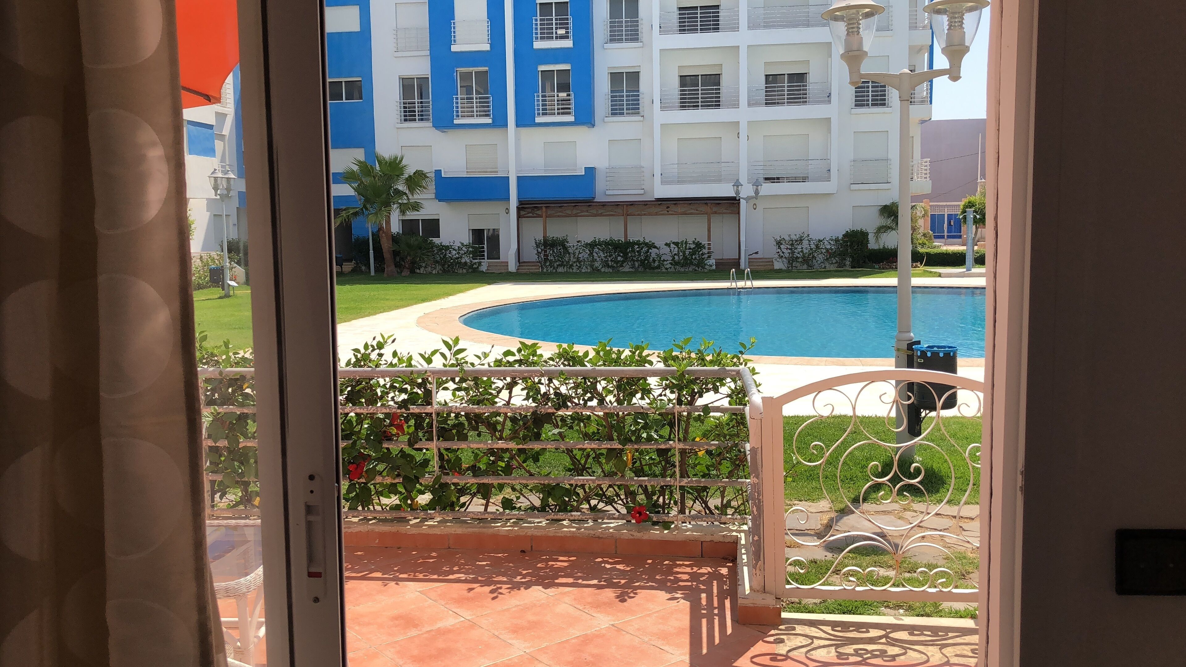 Vista de una piscina con fuente de agua azul, rodeada por un jardín cuidado y un edificio con paredes exteriores azules y blancas.