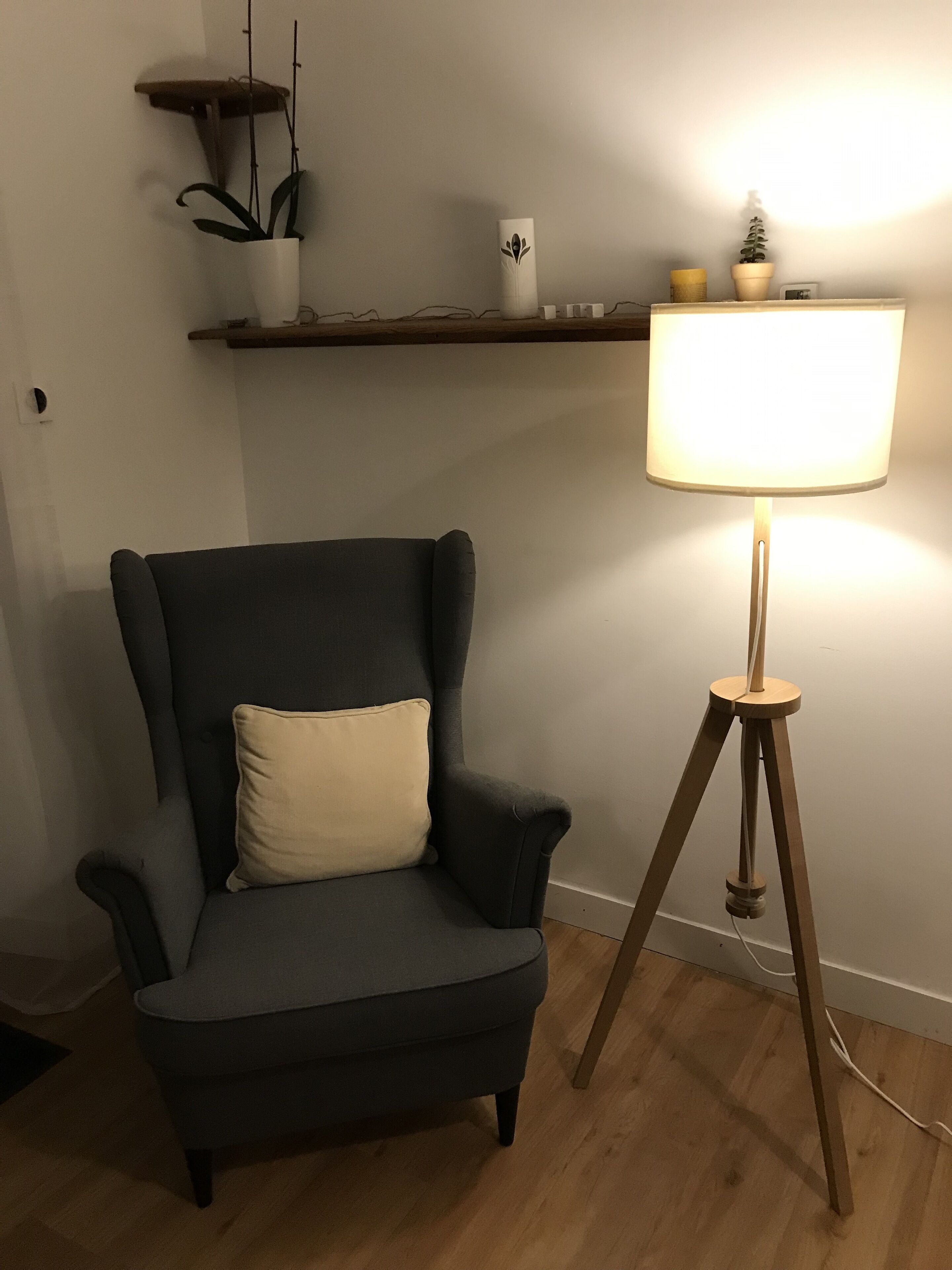 A dark grey armchair with a beige cushion, a wooden floor lamp with a white shade, and a wooden shelf above the chair.