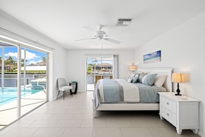A bedroom with a bed, nightstand, and lamp, a chair, a small table, and a view of a pool and outdoor area. - SEASIDE VILLA KEY BISCAYNE  (Cape Coral)