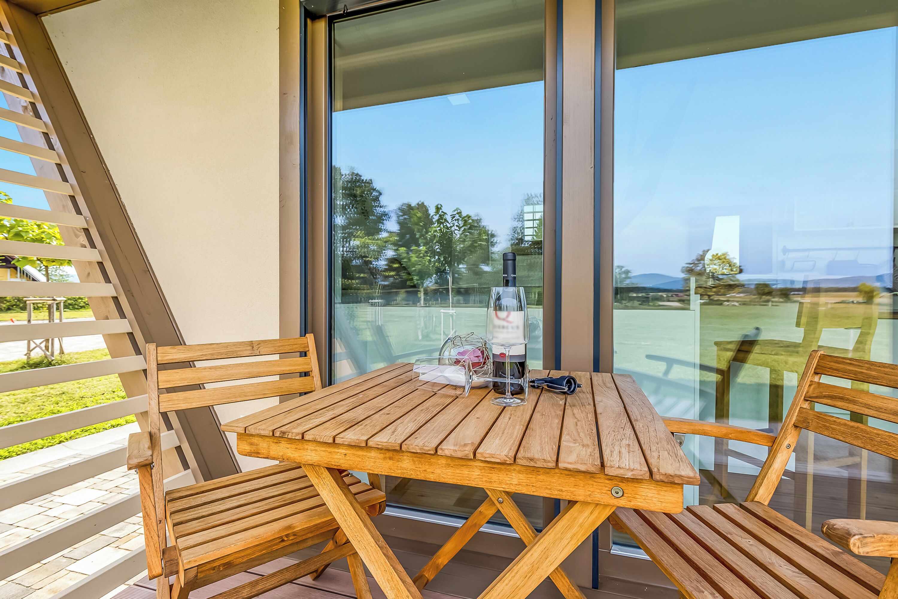 A wooden table and chairs on a balcony with a glass door, a bottle, and a wine glass.