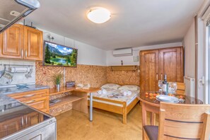 A compact room with a bed, a small table, a TV mounted on the wall, and a kitchenette area.