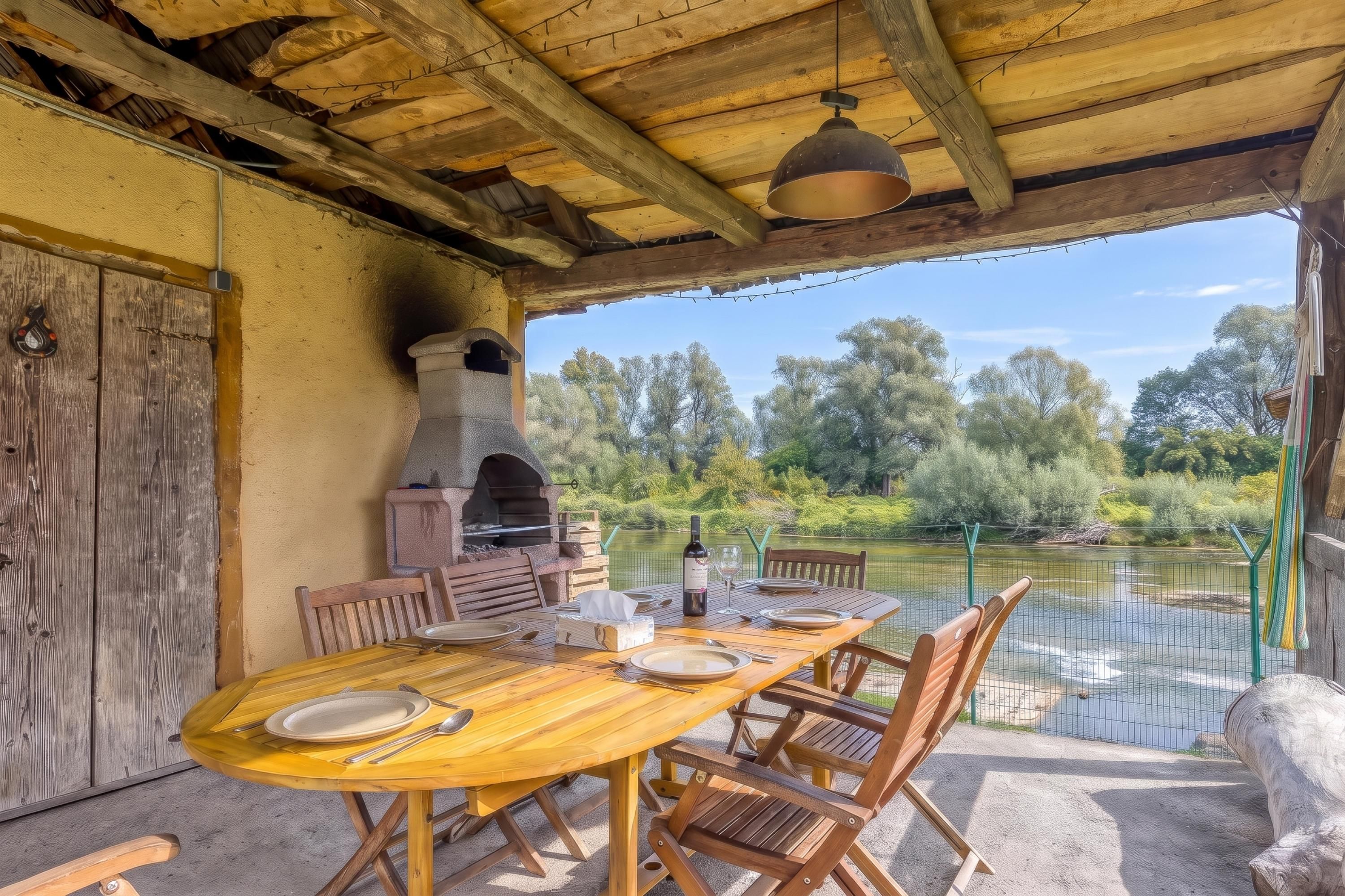 An outdoor dining area with a wooden table and chairs, a brick oven, and a view of a lake.