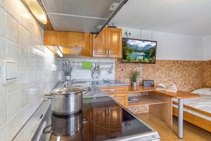 A compact kitchen with a stove, sink, and wooden cabinets. A flat-screen TV is mounted on the wall displaying a scenic landscape.