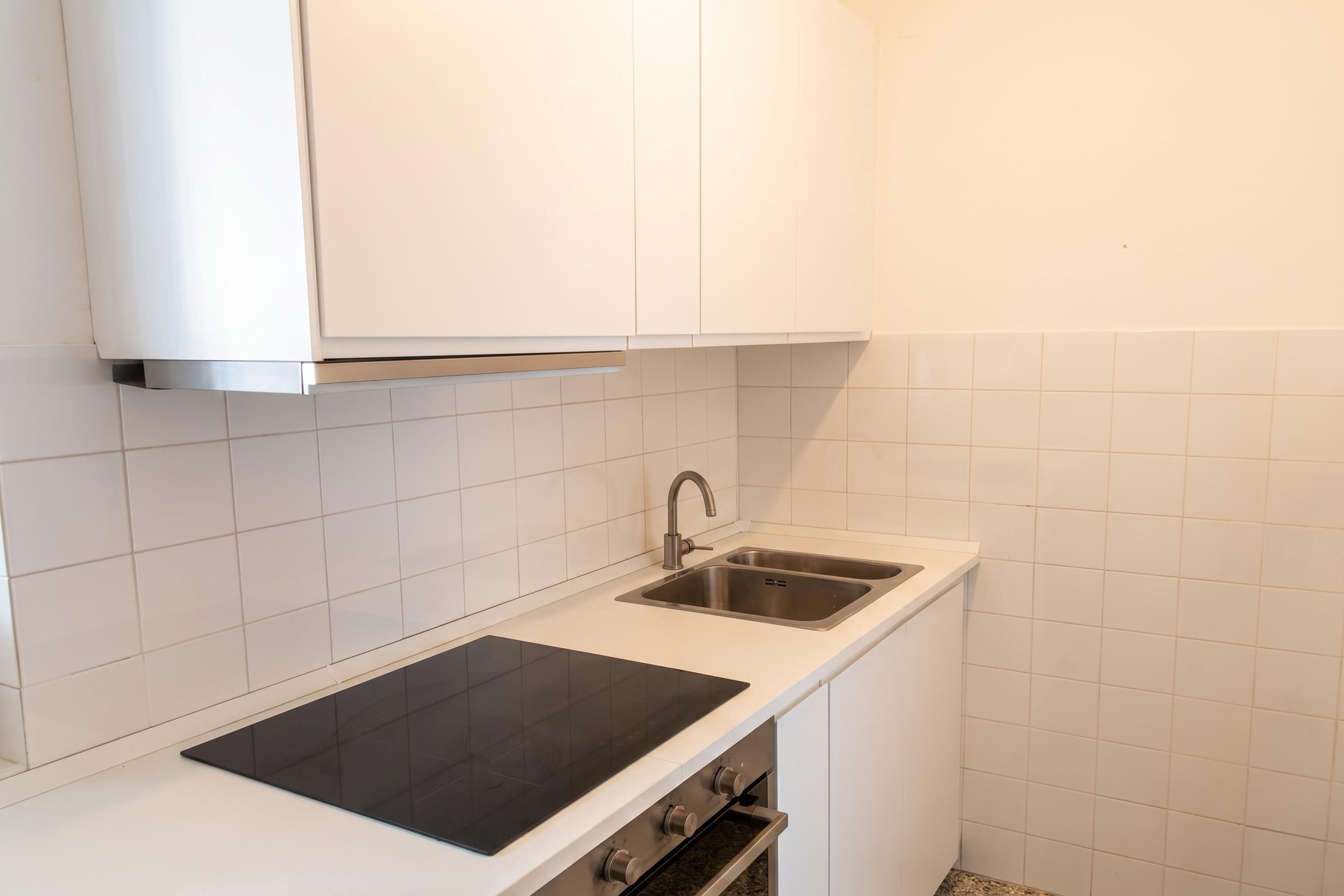 Une cuisine avec des meubles blancs, une plaque de cuisson à induction noire, un évier en acier inoxydable et un four encastré.