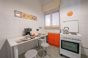 A compact kitchen with white tiled walls, a white countertop, a stove, a sink, and a window with lace curtains.