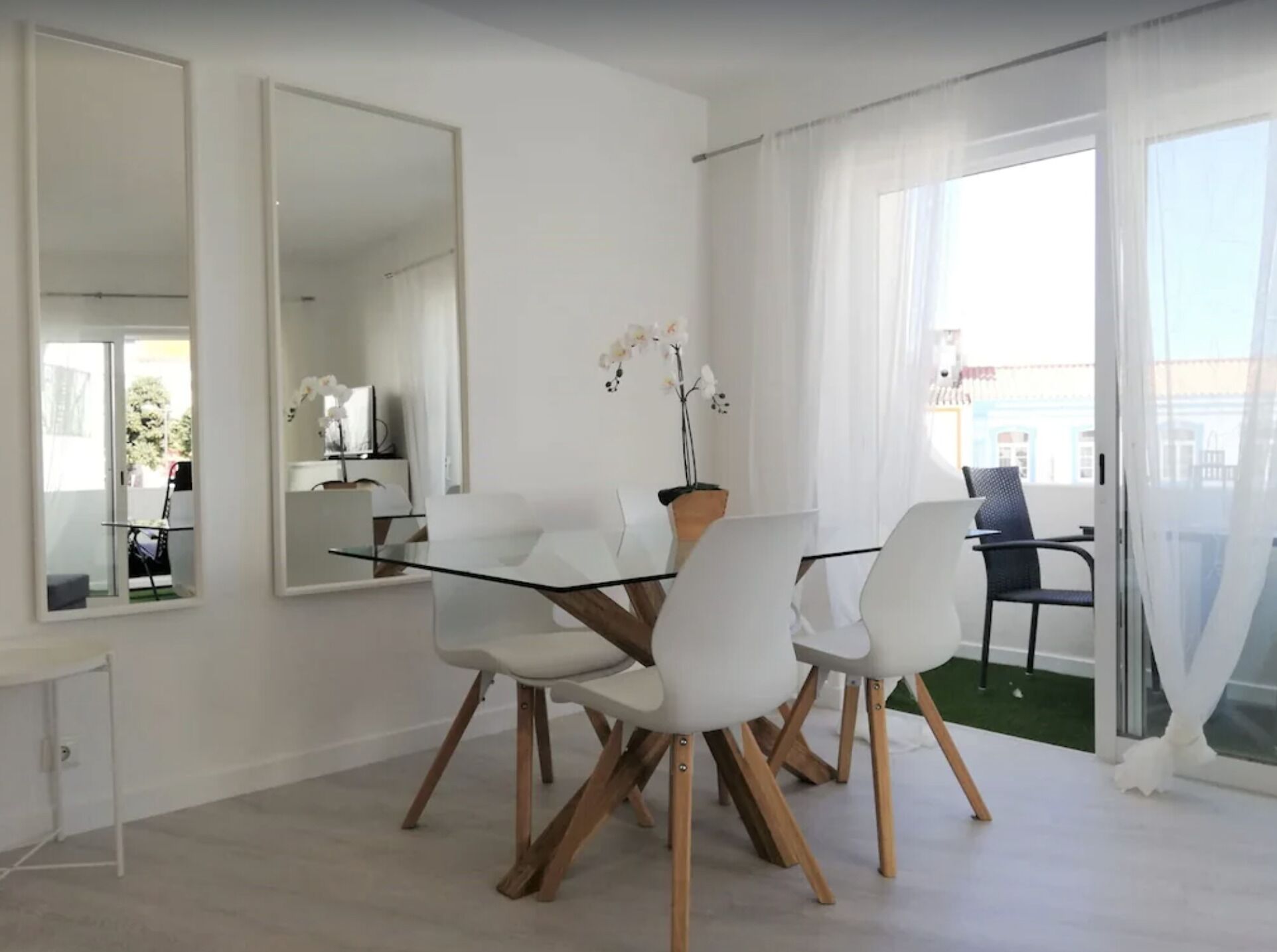 A modern dining area with a glass table and white chairs, a large mirror, and a view of the outdoors.