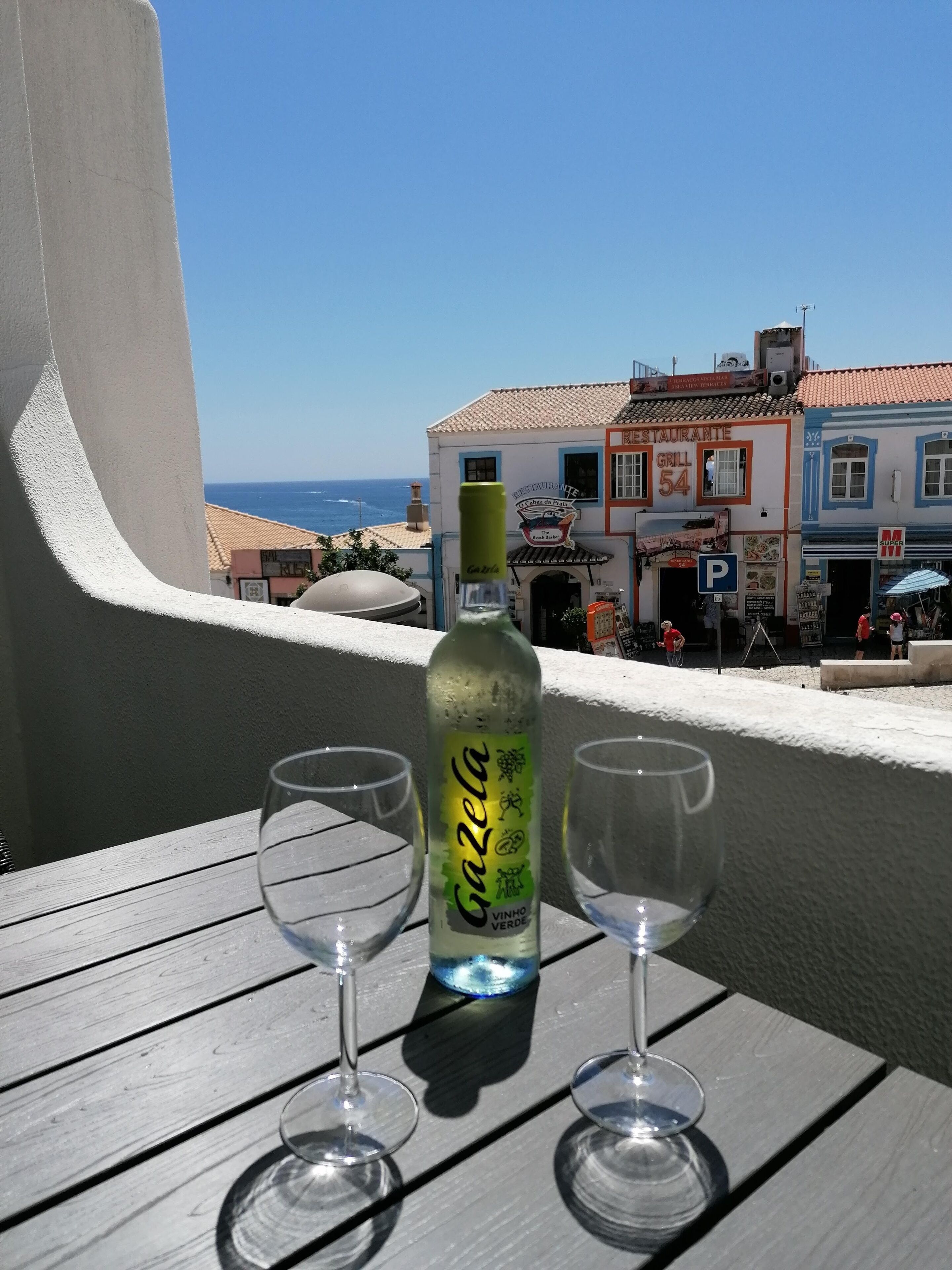 Un balcón con una botella de vino Gazela y dos copas de vino vacías, con vistas a una ciudad costera con edificios y cielo despejado.