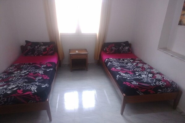 Une chambre avec deux lits simples, une petite table de chevet en bois et une fenêtre avec des rideaux.