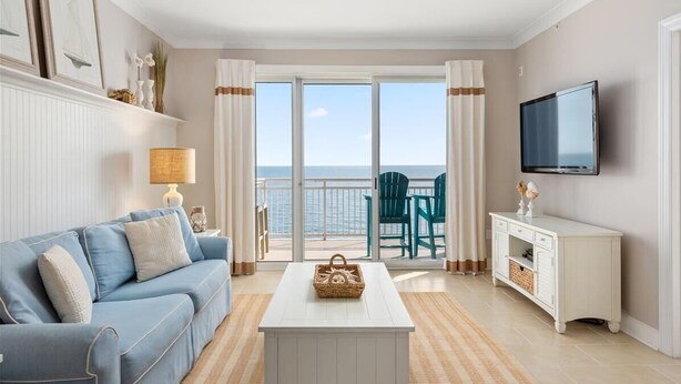 Una sala de estar con un sofá azul, una mesa de centro blanca, un televisor y vista al mar a través de un ventanal.