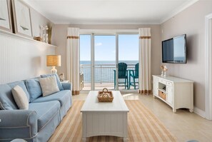 Ein Wohnzimmer mit einem blauen Sofa, einem weißen Couchtisch, einem Fernseher und Meerblick durch eine Glastür.