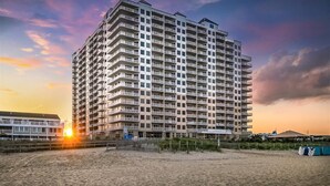 Un hotel de altura con varios balcones, cerca de una playa con un atardecer de fondo.