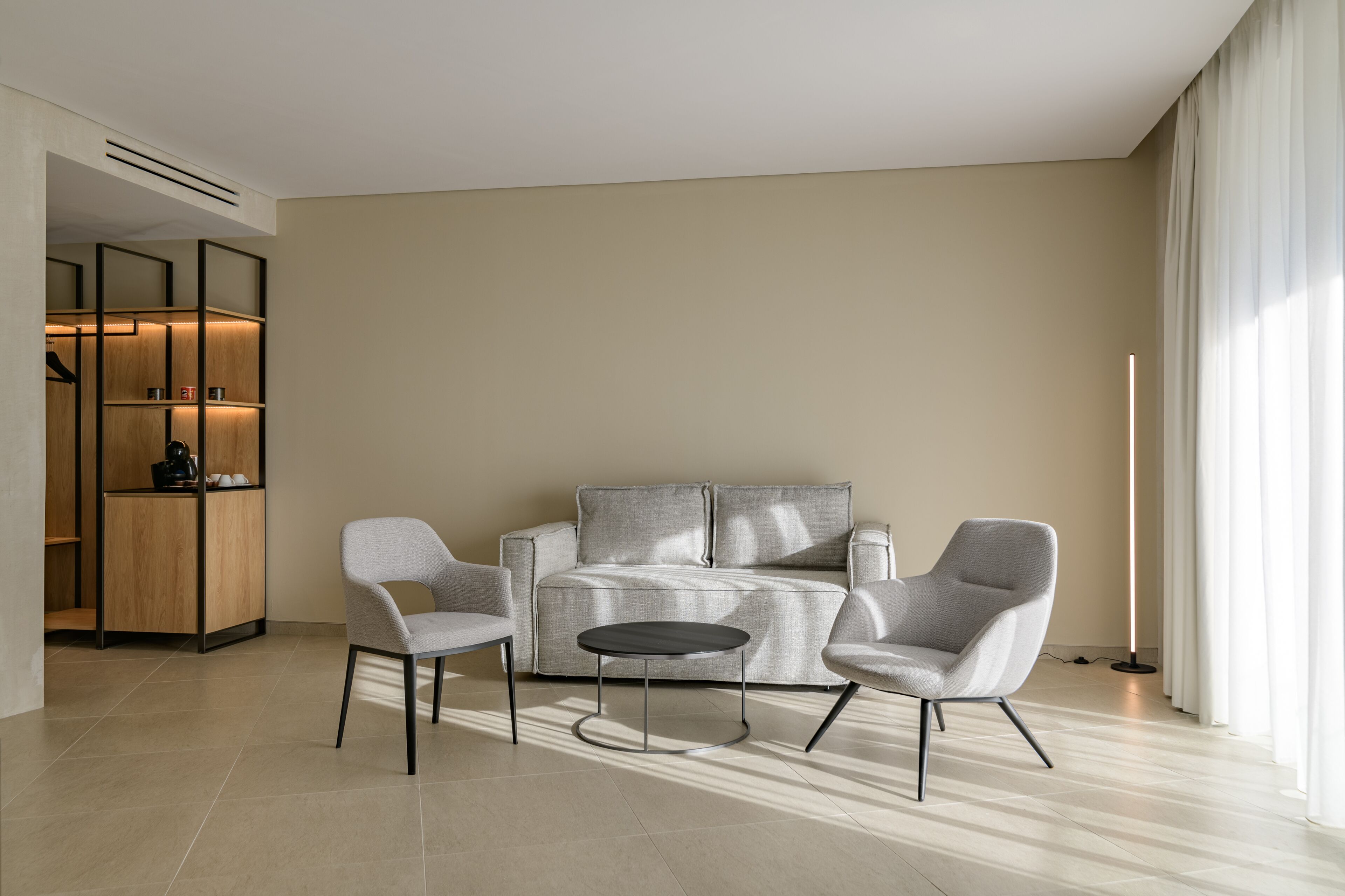 A modern living room with a sofa, two armchairs, and a small round table. There is a built-in shelving unit with a coffee machine and decorative items. The room has a large window with curtains and a floor lamp.