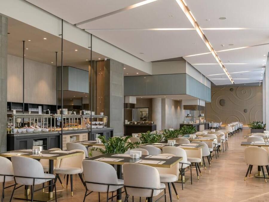 Un espace repas moderne avec de longues tables et des chaises, une cuisine en arrière-plan et un mur décoratif orné de motifs circulaires.