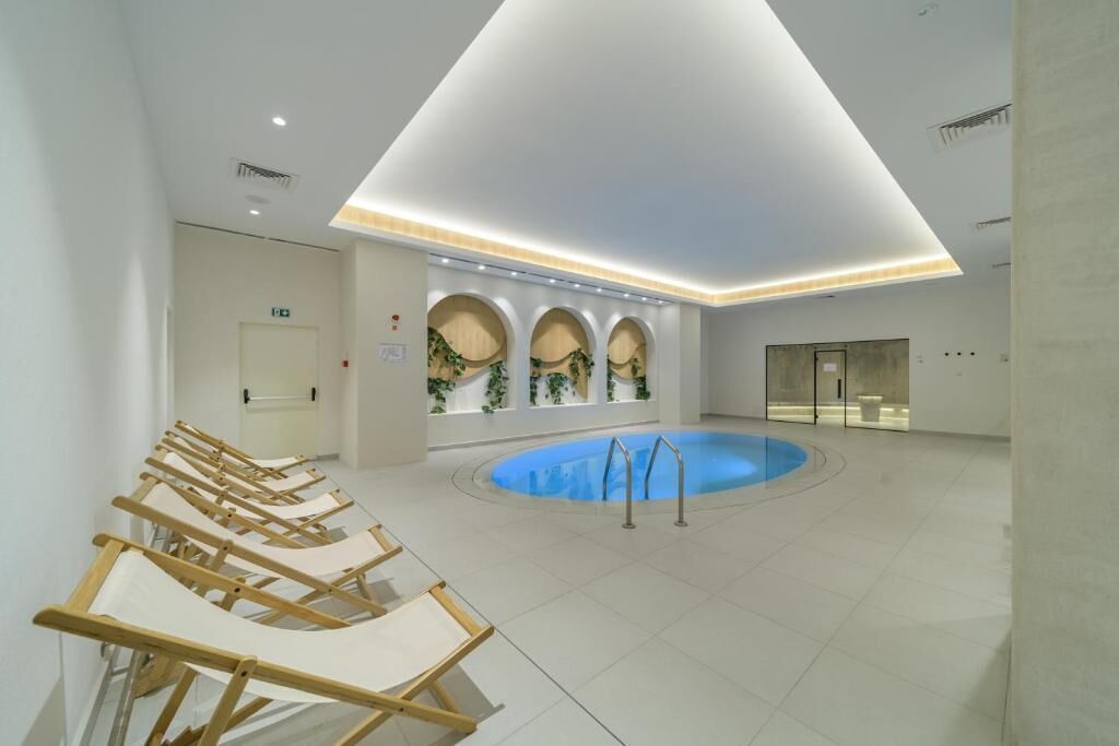 Un área de piscina interior moderna con una piscina central, sillas de descanso e iluminación empotrada en el techo.