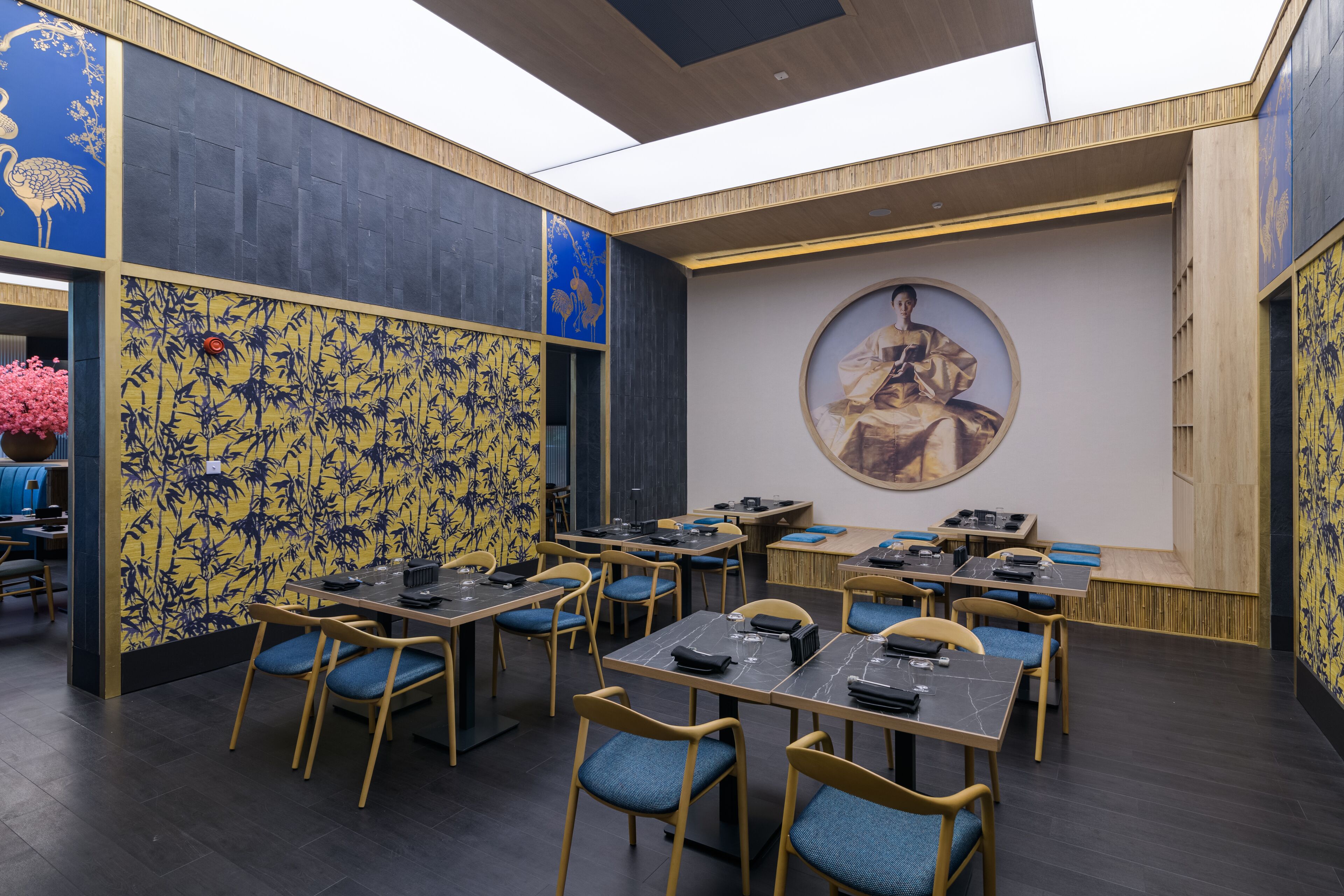 A restaurant with multiple tables and chairs, featuring a large mural on the wall and a ceiling with skylights.