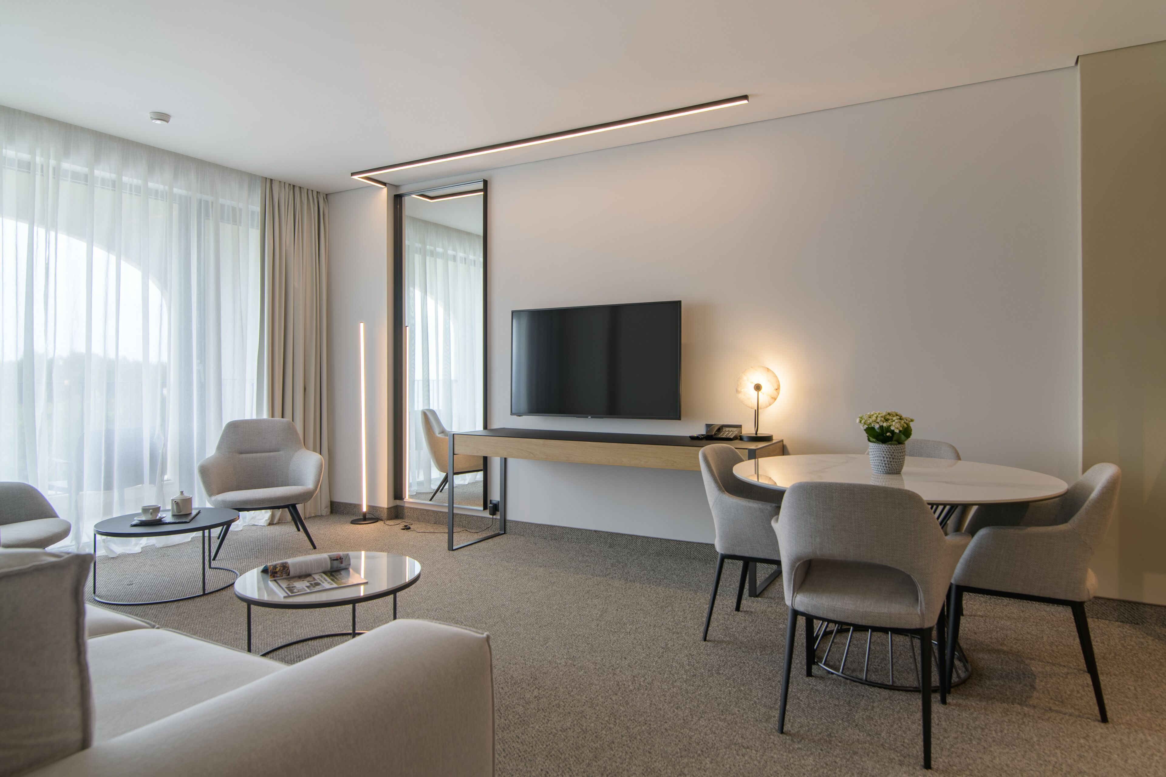 A modern hotel room with a flat-screen TV, a round dining table, and several chairs. There is a sofa and a small coffee table with a magazine.