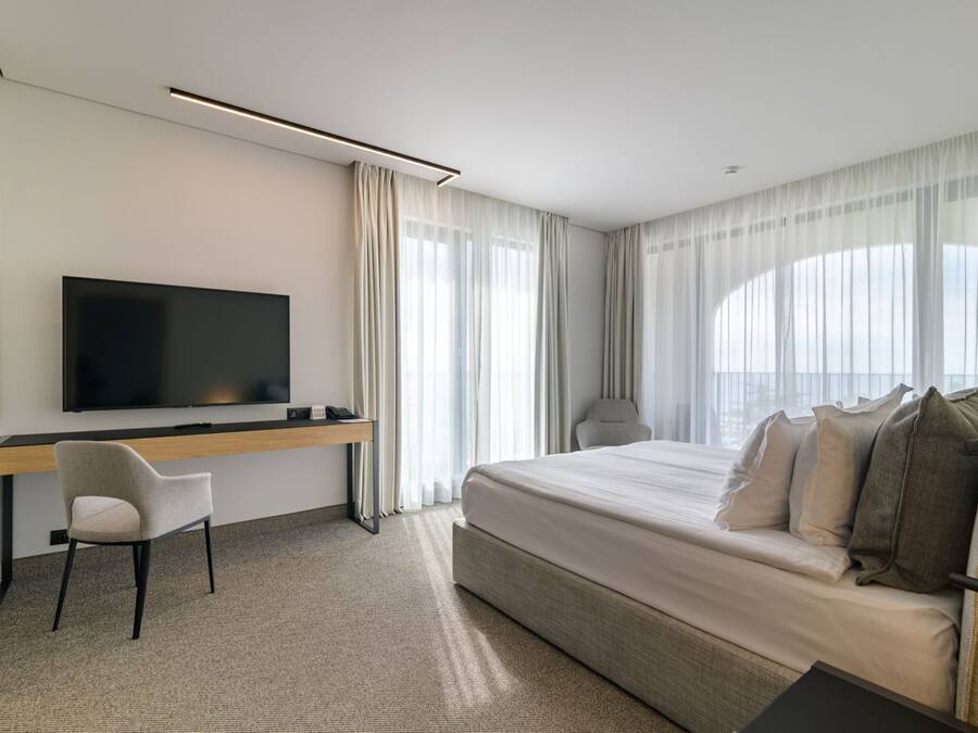 Una habitación de hotel moderna con una cama grande, un escritorio con una silla, un televisor de pantalla plana y ventanas amplias con cortinas traslúcidas.