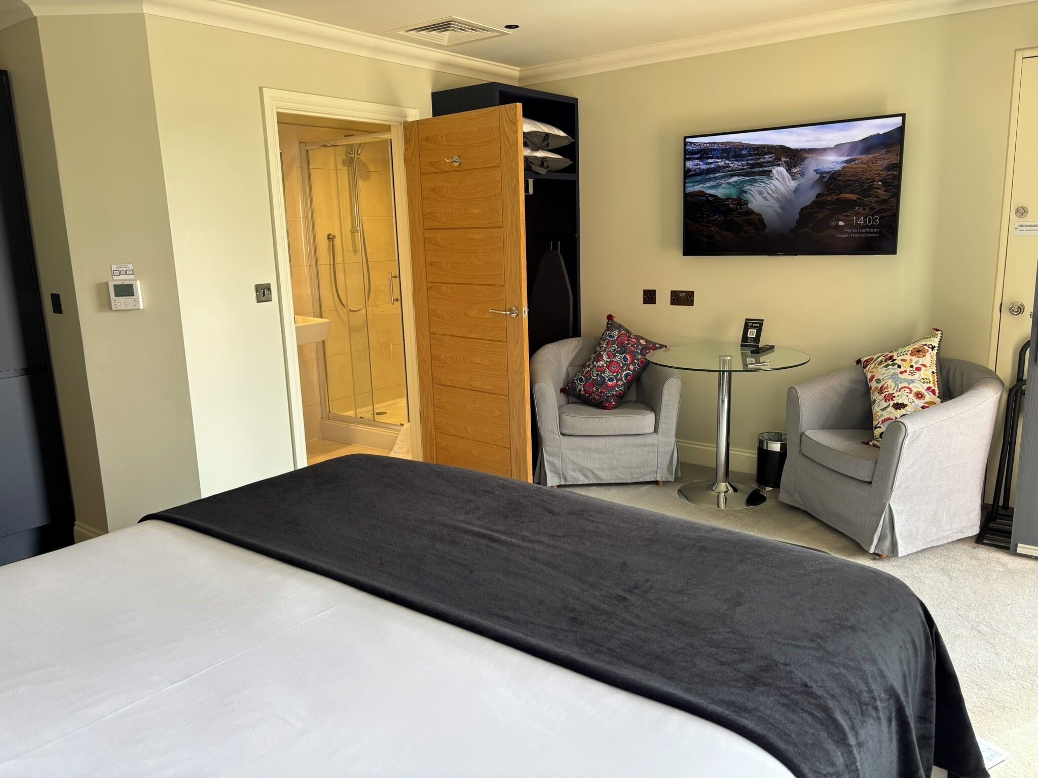 Quarto de hotel com cama, área de estar com duas cadeiras, uma mesinha e televisão fixada na parede, além de chuveiro.