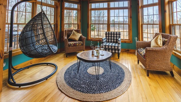 Une pièce chaleureuse avec un fauteuil suspendu, deux fauteuils, une table ronde et de grandes fenêtres donnant sur l’extérieur.