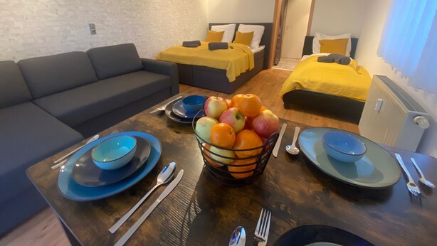 A modern living room with a sofa, a dining table with plates, bowls, and a fruit bowl. There are two beds with yellow bedding in the background.