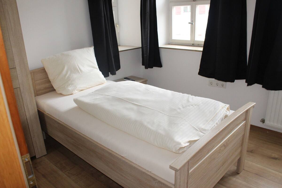 Una cama individual con estructura de madera y ropa de cama blanca en una habitación con ventana y cortinas negras.