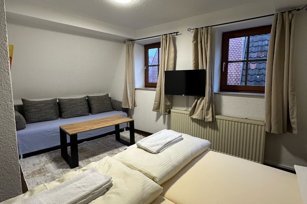 Una habitación con cama, sofá, mesa, televisor y ventana con cortinas.