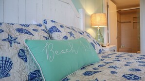 Een slaapkamer met een bed met een blauw-wit schelpenmotief, een groen kussen met de tekst 'Beach Life' en een lamp op een nachtkastje.