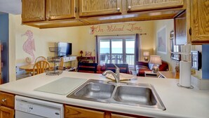 A kitchen with wooden cabinets, a double sink, a dining table with chairs, a television, and a view of the ocean through a sliding door. - 2 Bedroom Oceanfront Condo with Beautiful Views (Surf City)