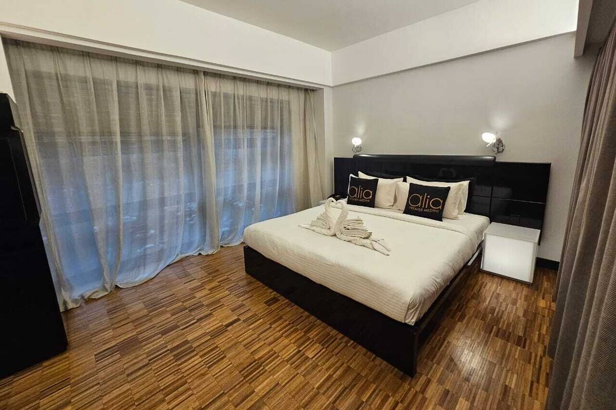 A modern hotel room with a large bed, a bedside table, and a window with sheer curtains.