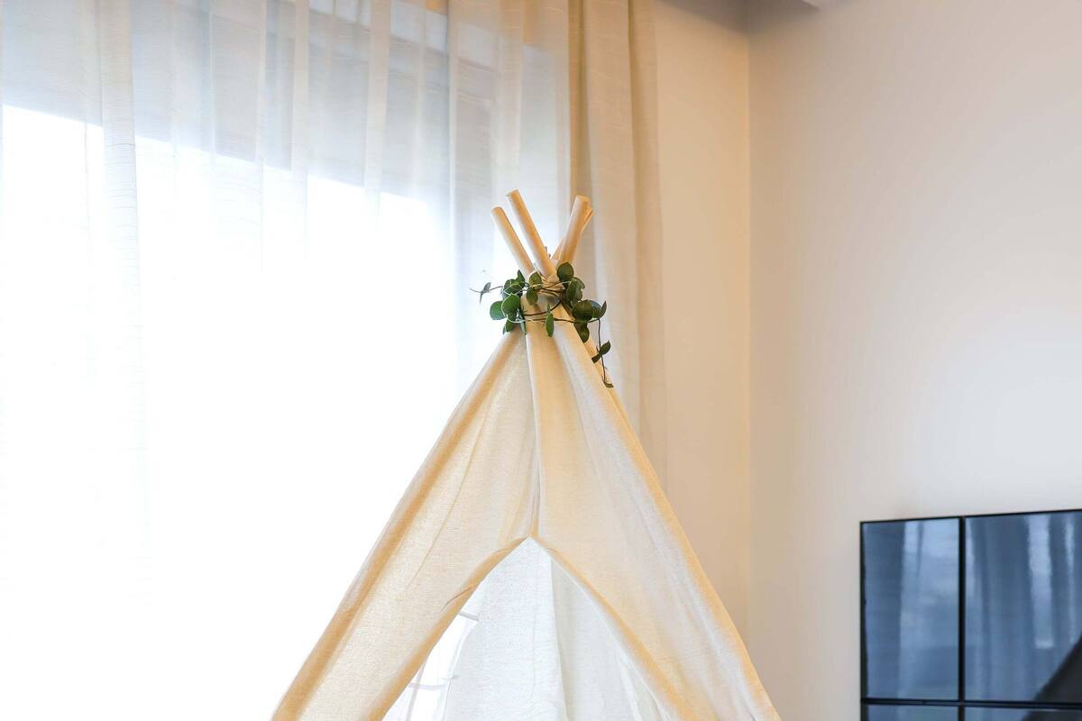 A cozy bedroom with a bed, a white furry rug, a tent-like structure with pillows, and a large window.